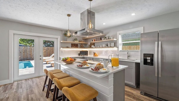 Cozinha moderna com ilha central, banquetas, geladeira de aço inoxidável e vista para um quintal com piscina.
