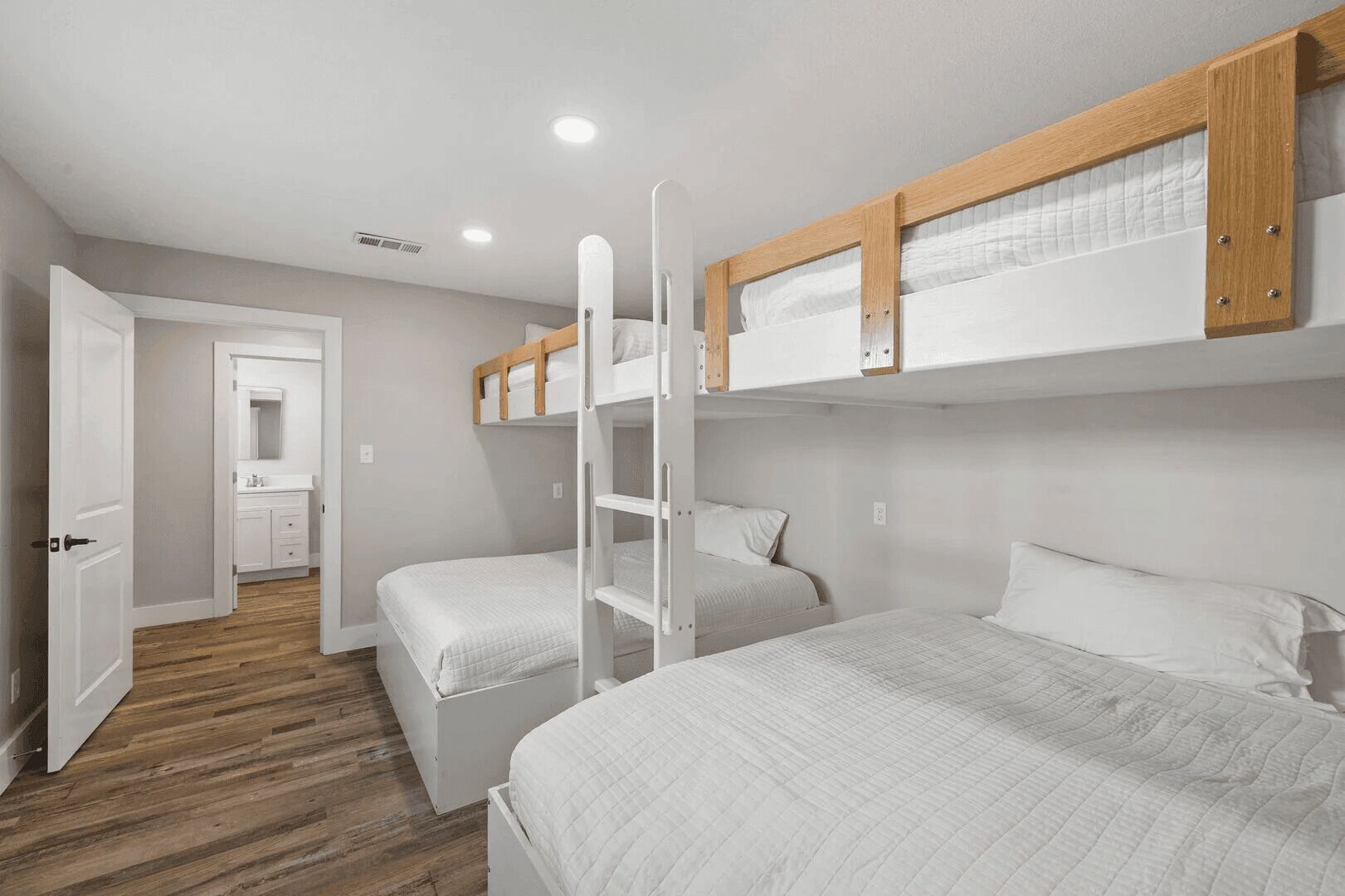 Un dormitorio con literas, suelo de madera y un baño visible a través de una puerta abierta.