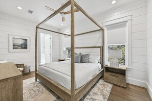 A bedroom with a four-poster bed, a wooden headboard, a bedside table with a lamp, a window with a view, and a framed picture on the wall. - Alibi 30A by Avantstay 6bdr, Private Pool, Steps to Beach (Panama City Beach)