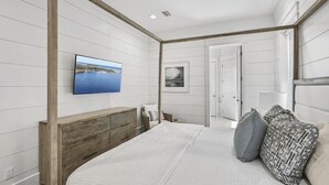 Un dormitorio con cabecera de madera, cama con ropa de cama blanca, ventilador de techo y televisor de pared mostrando un paisaje.