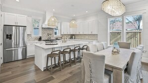 Una cocina moderna con armarios blancos, un refrigerador de acero inoxidable, una isla central con taburetes de bar y una araña.