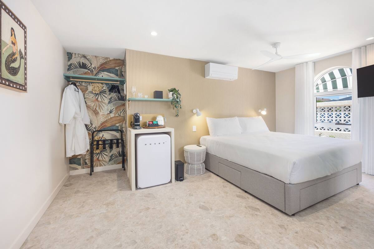 Une chambre d’hôtel moderne équipée d’un lit, d’un petit bureau avec une machine à café, d’un climatiseur mural et d’une plante décorative.