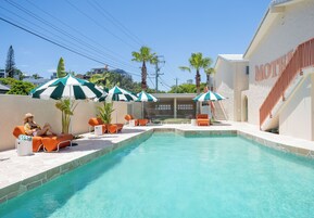Una piscina con sillones de descanso y sombrillas, una escalera que lleva a un motel, y un cielo despejado.