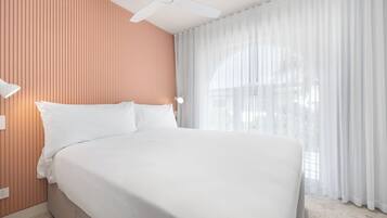 Una cama bien hecha en una habitación de hotel con ventilador de techo, una ventana con cortinas traslúcidas y una pared con listones de madera verticales.