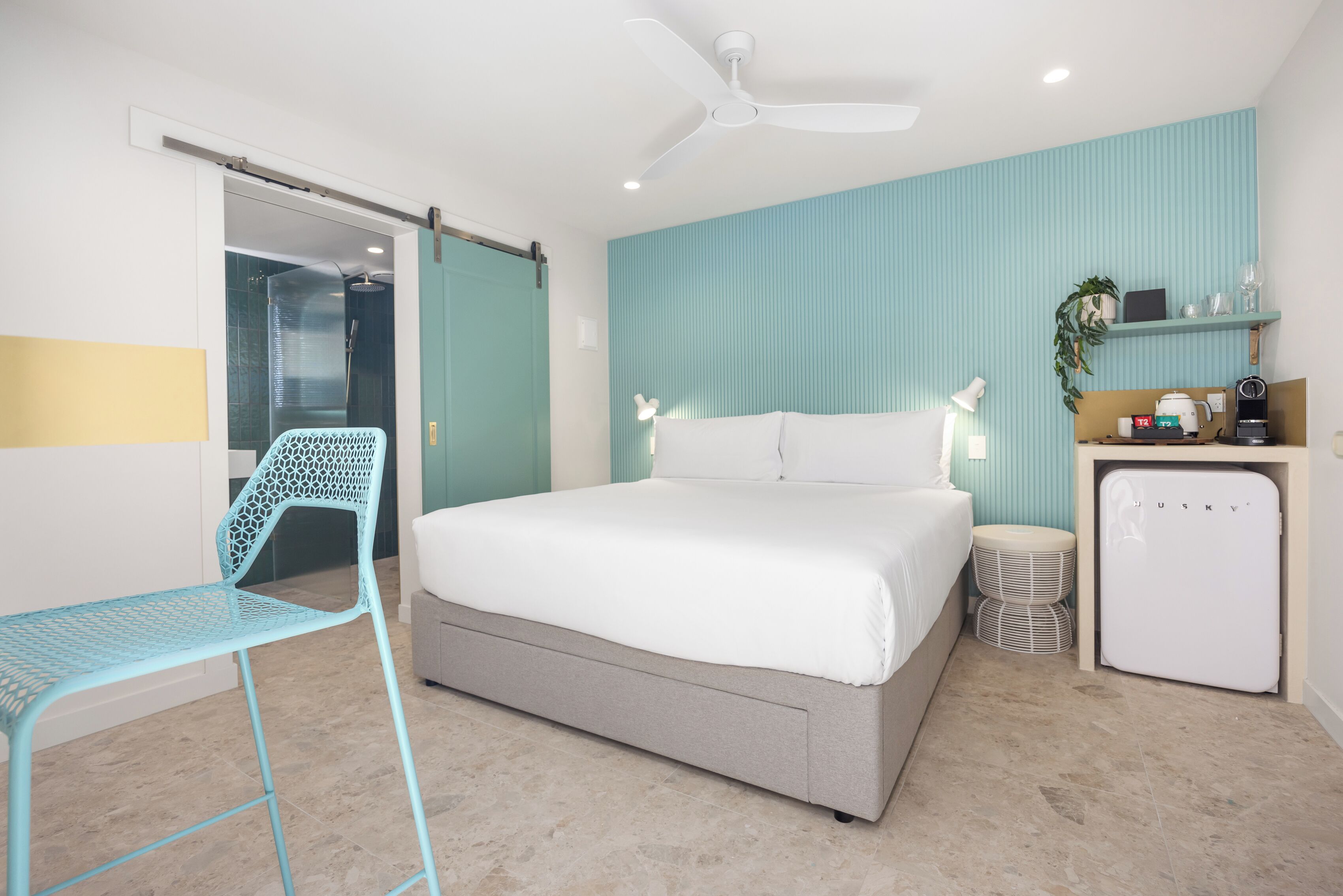 A bedroom with a bed, a desk, a chair, a ceiling fan, and a small refrigerator.