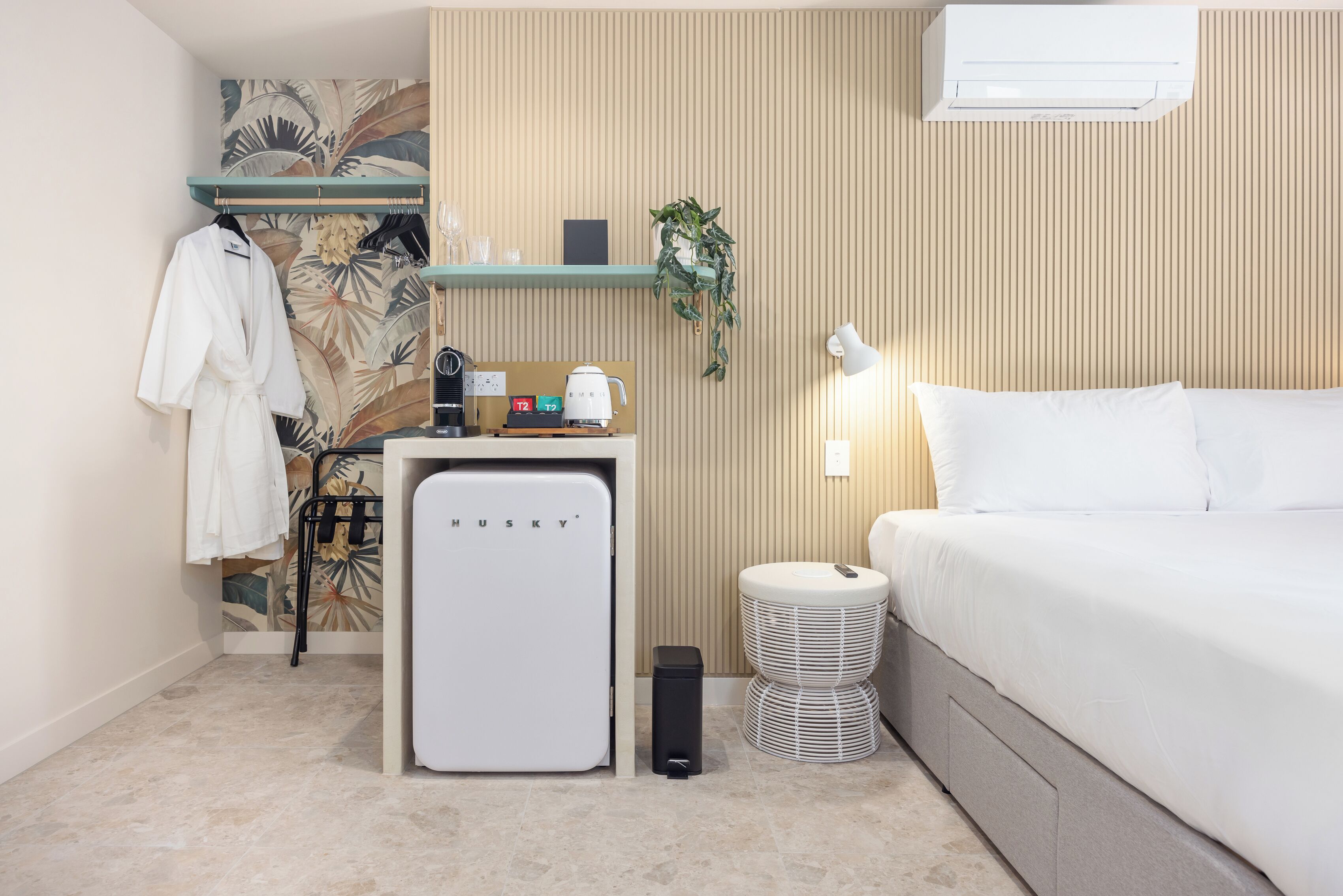 Habitación de hotel pequeña y funcional con cama, un escritorio, cafetera y aire acondicionado de pared.