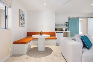 Una sala de estar moderna con un sofá blanco, cojines naranjas, una mesa redonda blanca y una zona de cocina al fondo.