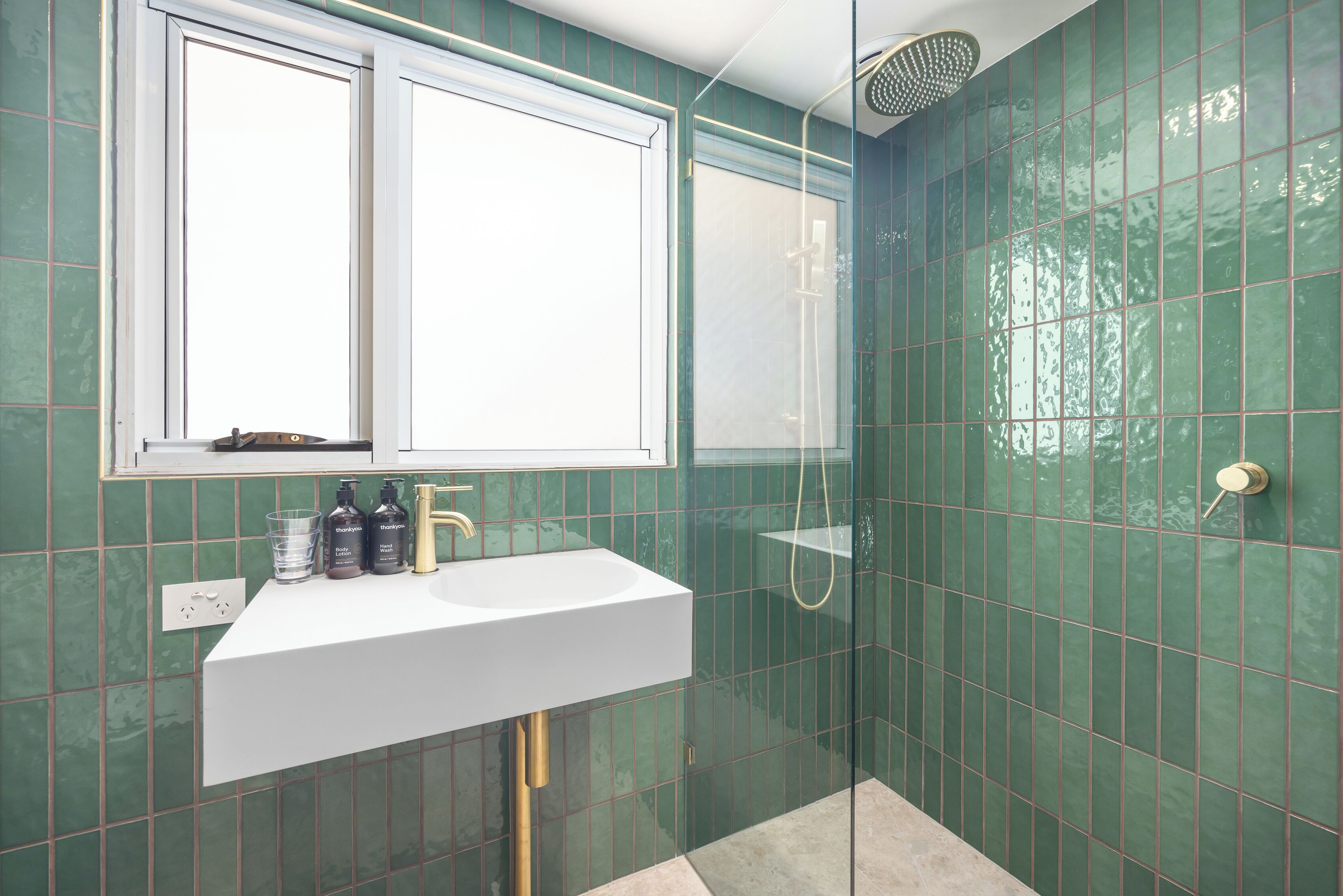 A bathroom with a white sink, green tiled walls, a window, and a shower area.
