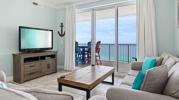 A living room with a television, wooden furniture, a sofa with teal cushions, and a balcony with a view of the ocean. - Beachfront Access, 2 Pools, Beach Service w/ Chairs Emerald Isle Resort 1308 (Panama City Beach)