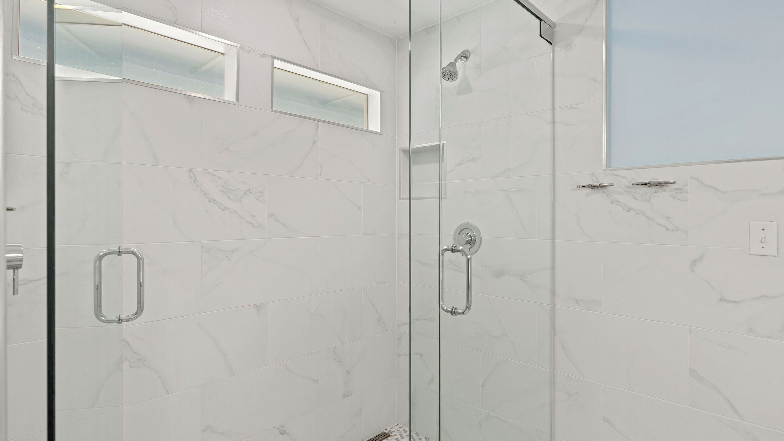 A modern bathroom with a glass shower enclosure, marble tiles, and a visible handrail.