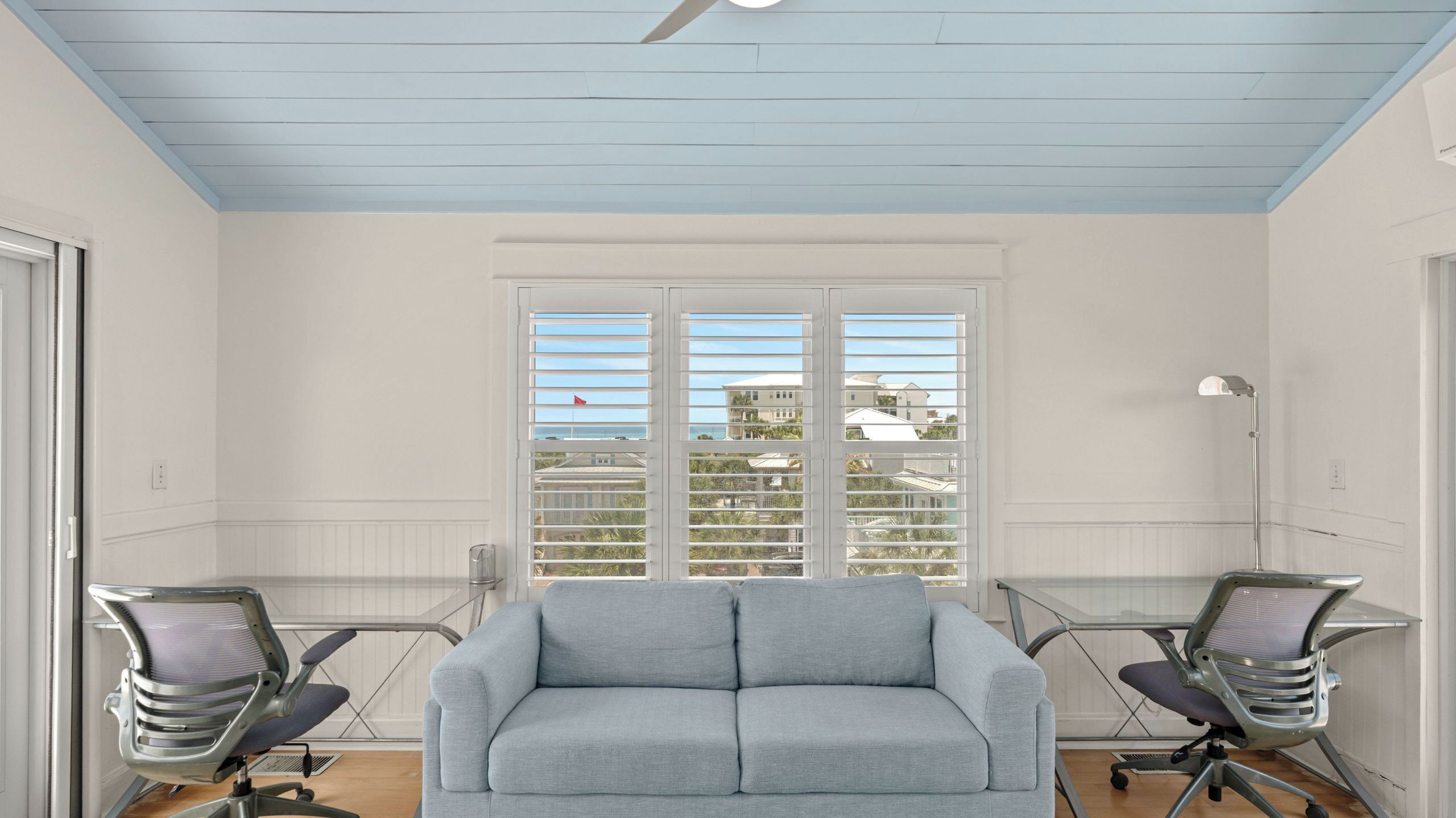A room with a light blue ceiling, a grey sofa, two office chairs, a desk, and a window with blinds.