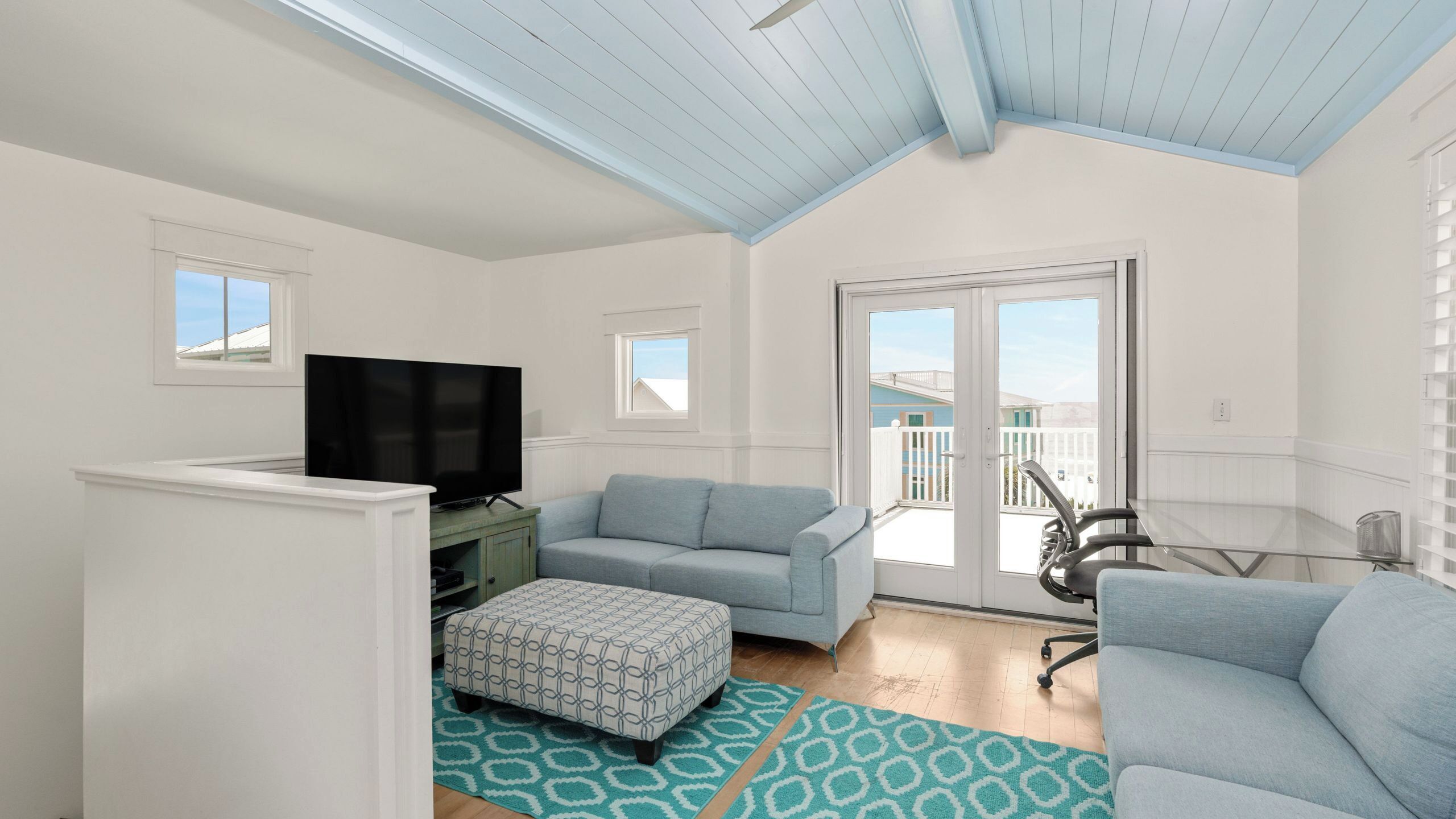 A living room with a TV, a sofa, an ottoman, a glass table, and a balcony door.