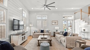A spacious living room with a large ceiling fan, a flat-screen TV mounted on the wall, and a staircase leading to an upper level. - 30A Aqua Vista by Avantstay Golf Cart Included, Pool Acess, Walk to Beach (Santa Rosa Beach)