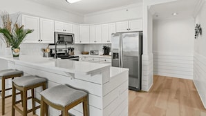 A modern kitchen with white cabinetry, a stainless steel refrigerator, and a central island with bar stools. - Coastal Hippy Chic by Avantstay Oceanfront, 2 Pools, Beach Chairs Incl (Panama City Beach)
