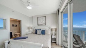 A bedroom with a large bed, a nightstand, a bedside table, and a view of the ocean through a balcony. - Coastal Hippy Chic by Avantstay Oceanfront, 2 Pools, Beach Chairs Incl (Panama City Beach)