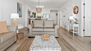 A modern living room with a beige sofa, a coffee table, and a kitchen in the background. - Coastal Hippy Chic by Avantstay Oceanfront, 2 Pools, Beach Chairs Incl (Panama City Beach)