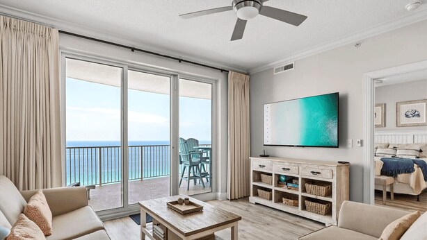A spacious living room with a large sliding glass door leading to a balcony with a view of the ocean. There is a flat-screen TV mounted on the wall, a wooden coffee table, and beige sofas.