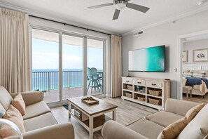 A spacious living room with a large sliding glass door leading to a balcony with a view of the ocean. There is a flat-screen TV mounted on the wall, a wooden coffee table, and beige sofas. - Manta Ray Villa by Avantstay Gulf Front + 2 Pools (Panama City Beach)