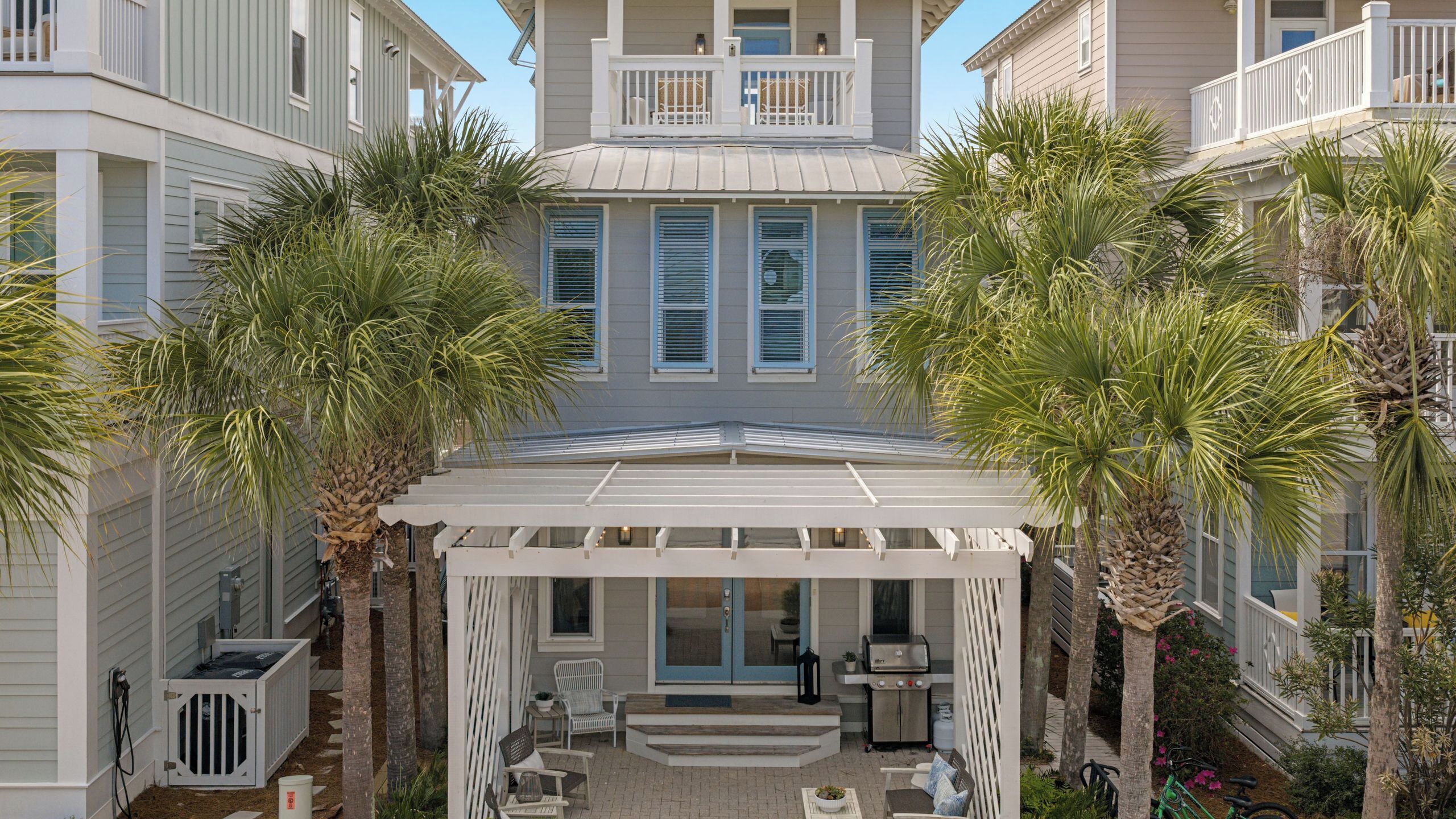 A covered outdoor seating area with a pergola, palm trees, and a building with a balcony.