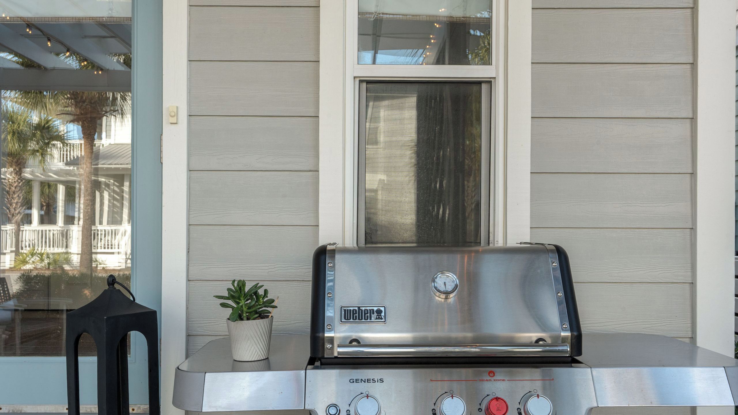 A Weber Genesis gas grill with a digital display, positioned in front of a house with light-colored siding and a window above it.