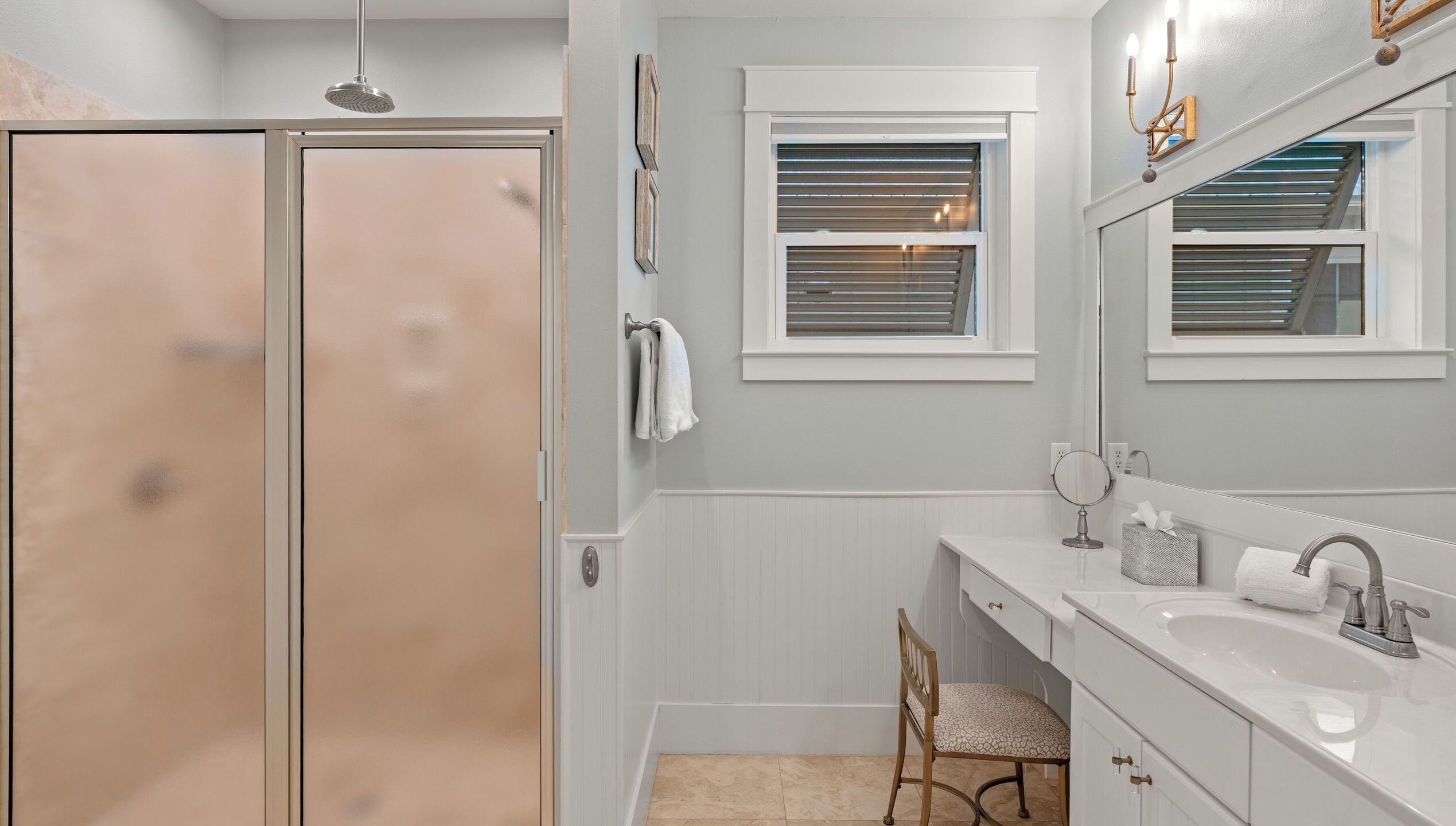 A bathroom with a shower, vanity, mirror, and a window with blinds.