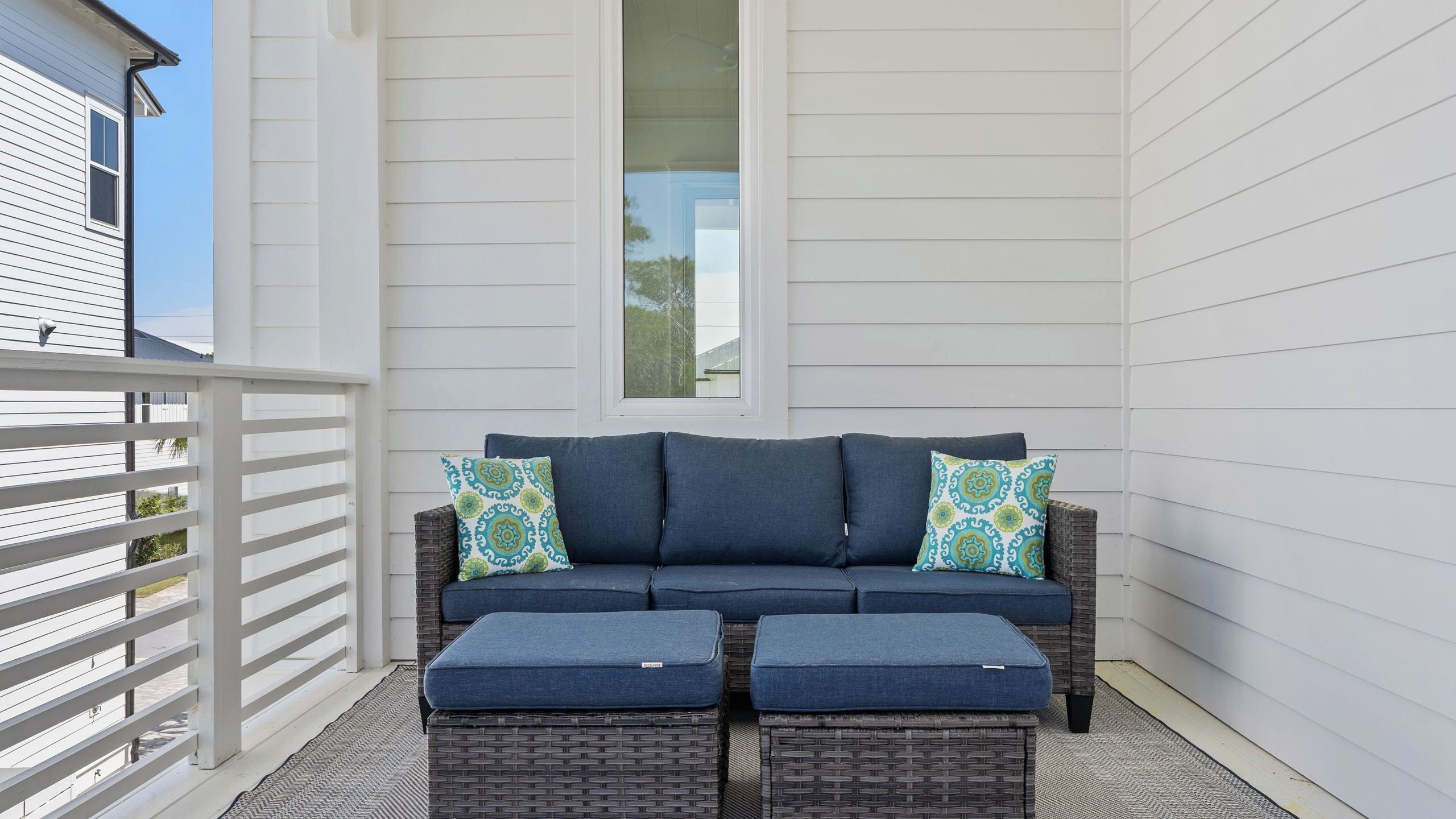 A balcony with a wicker sofa and ottomans, a white railing, and a window.