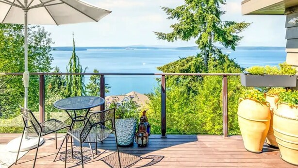 Eine Terrasse mit Tisch und Stühlen, einem weißen Sonnenschirm und Kübelpflanzen.