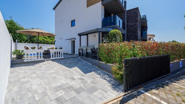 Una casa moderna a due piani con patio lastricato, area salotto esterna e ingresso recintato.