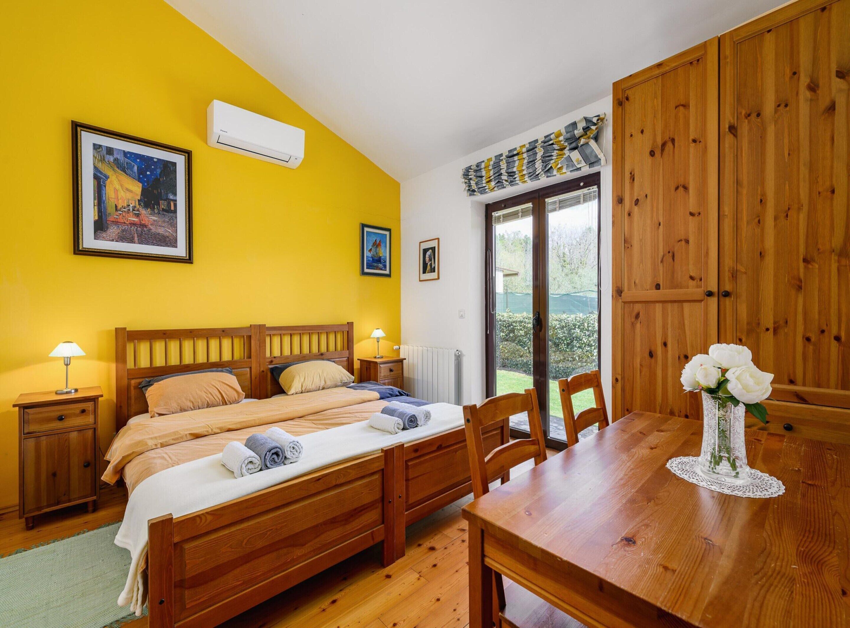 A bedroom with a bed, bedside tables, a wooden dining table, chairs, a radiator, a window with curtains, and a view of greenery outside.