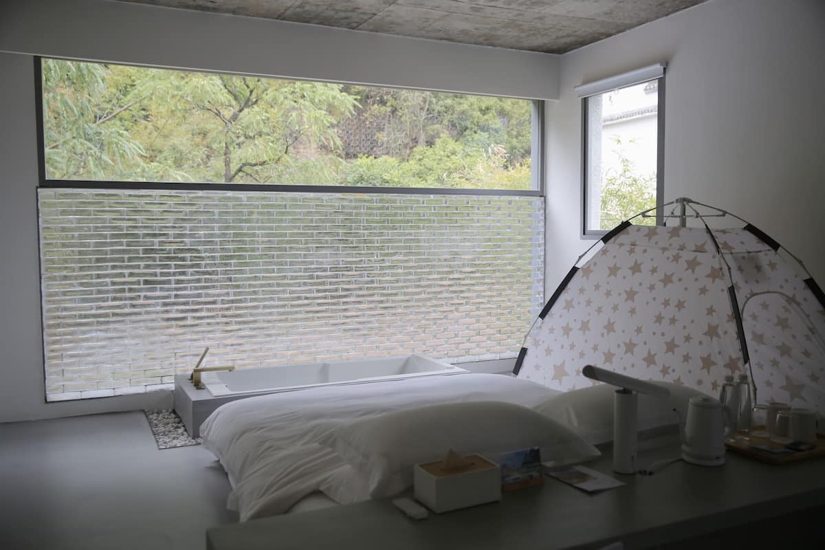 A bedroom with a large window, a bed, a bedside table with a lamp, and a star-patterned tent-like structure.