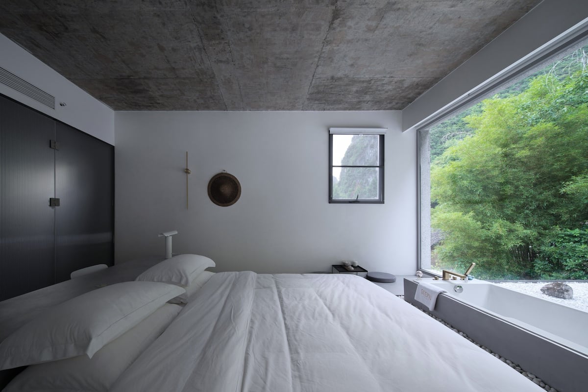A modern bedroom with a large bed, a window with a view of greenery, a wall-mounted clock, and a small bedside table with a lamp.