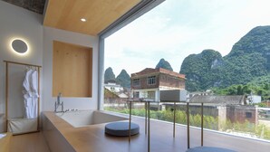 A modern bathroom with a large bathtub, a wooden vanity, and a view of a mountainous landscape through large windows. - Yangshuo Geemu Building House (Guilin)
