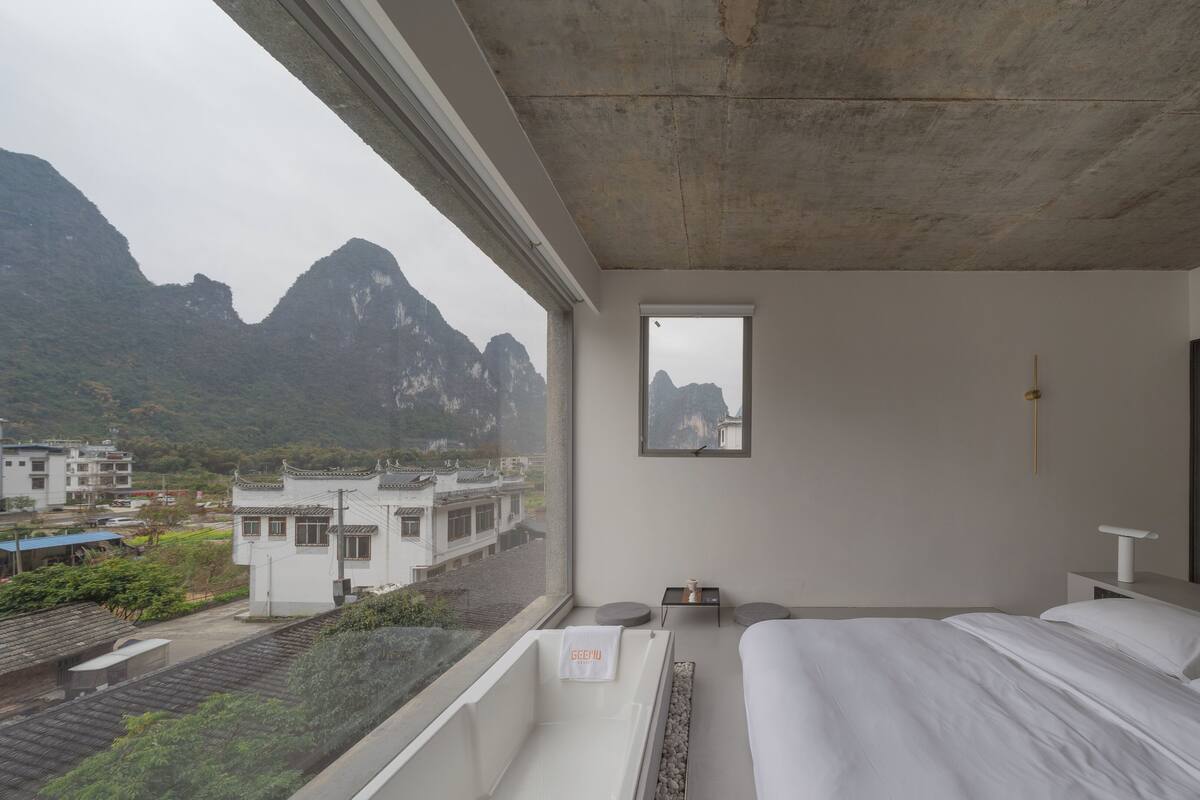 A hotel room with a large window, a bed, a bedside table, and a view of mountains and buildings.
