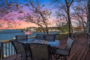 Une terrasse dotée de mobilier d’extérieur, donnant sur un lac, avec un drapeau américain et une vue sur une côte lointaine parsemée de bâtiments.
