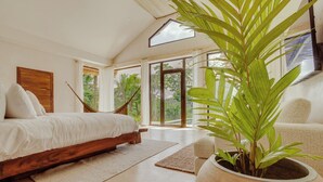 Une chambre avec un lit en bois, une grande plante en pot et une vue sur la verdure à l’extérieur.