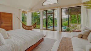 Une chambre avec un grand lit, un hamac, un canapé et une vue sur une piscine et la verdure.