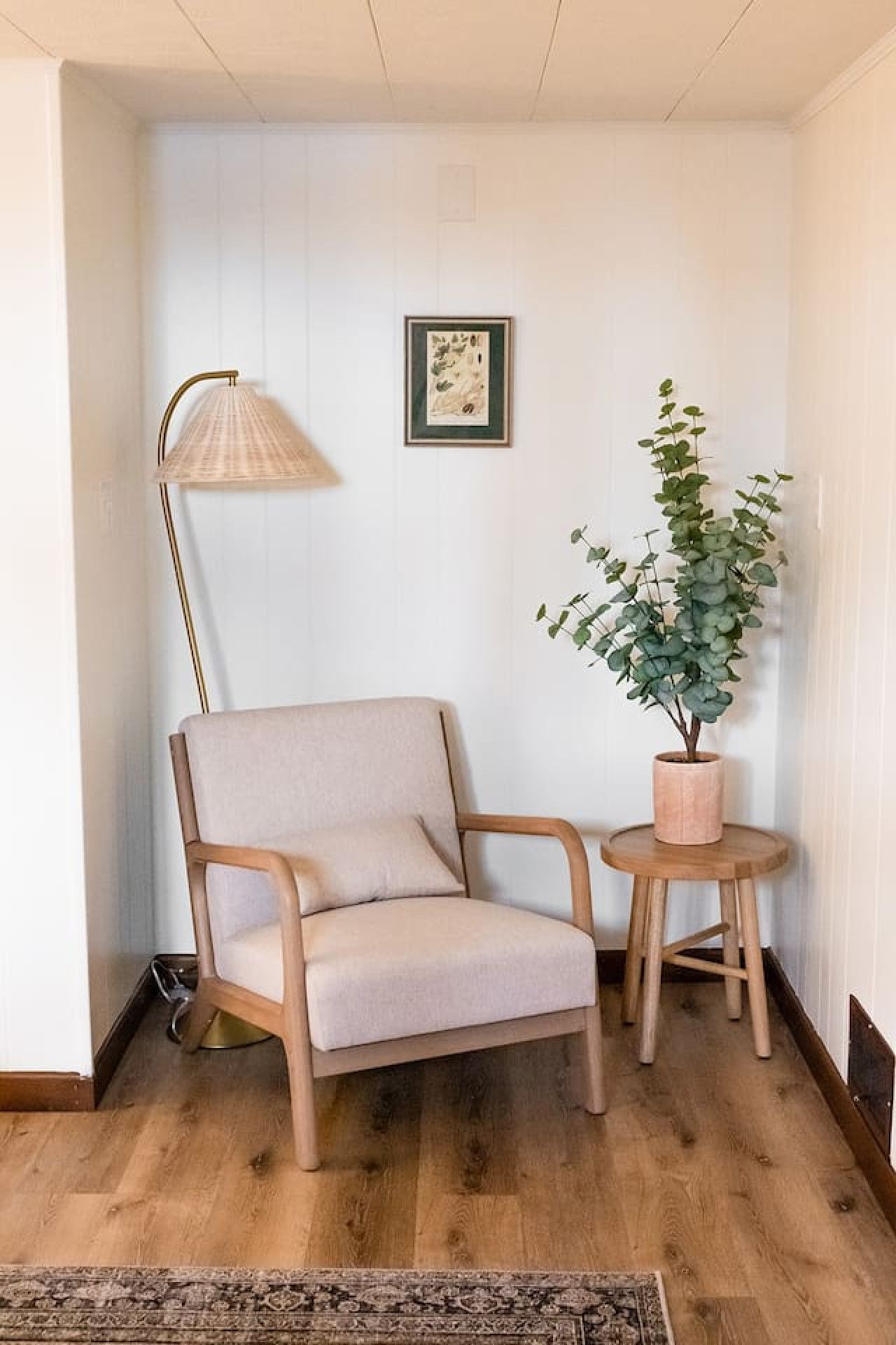 Habitación en esquina con piso de madera, un sillón beige, un taburete de madera, una lámpara de pie y una planta en maceta.