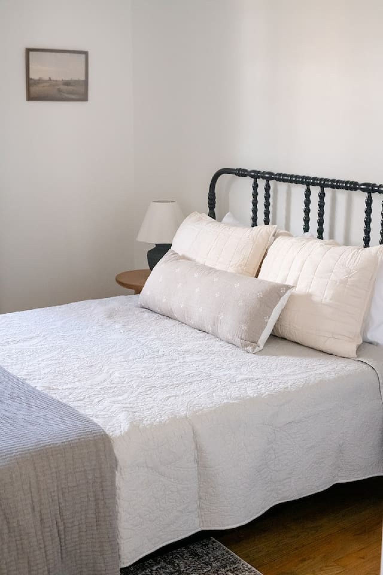 Ein ordentlich bezogenes Bett mit weißer Steppdecke und Kissen, ein schwarzes Metall-Kopfteil, ein Nachttisch mit Lampe und ein gerahmtes Bild an der Wand.