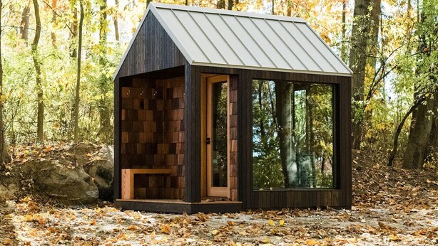 Une petite cabane dotée d’un toit en pente et d’une porte en verre, entourée d’une forêt où les feuilles sont tombées.