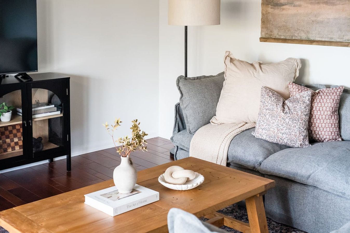 Un soggiorno con un divano grigio, un tavolino in legno, un vaso bianco con fiori secchi e una lampada.