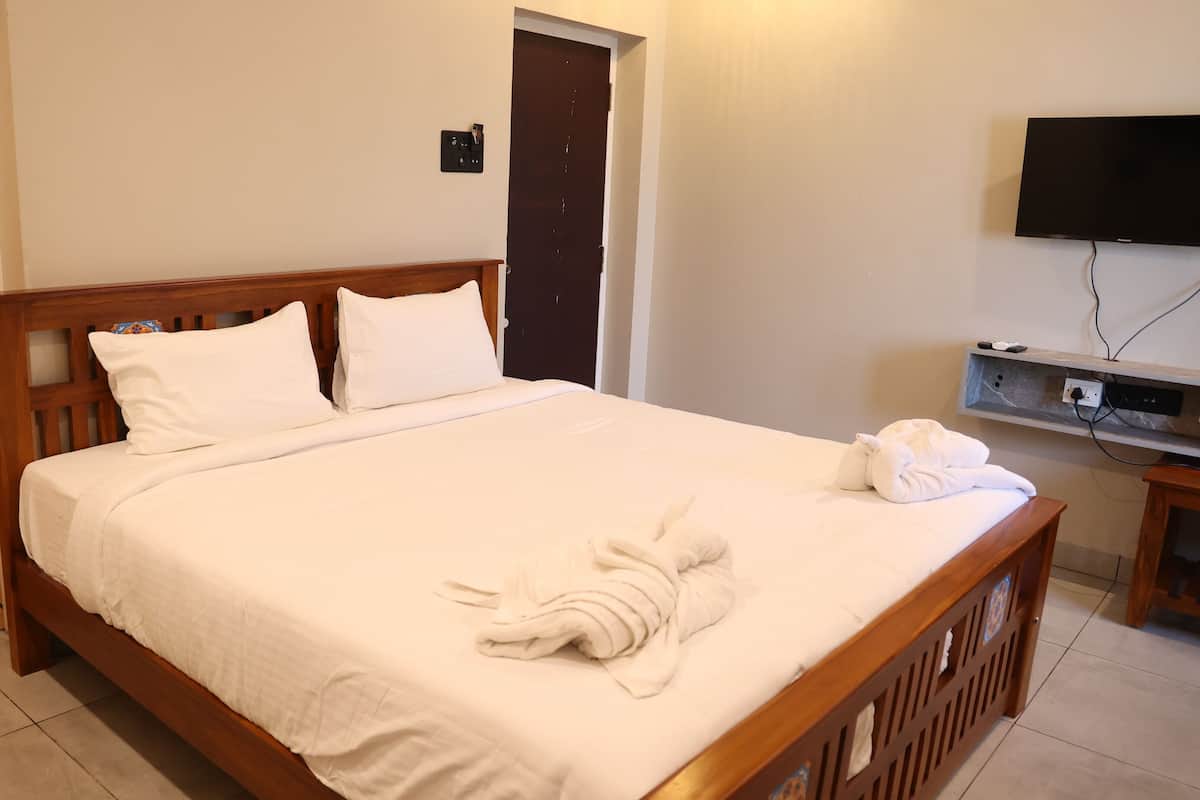 Een opgemaakt bed met witte lakens, een houten hoofdbord, een aan de muur bevestigde televisie, en een deur op de achtergrond.