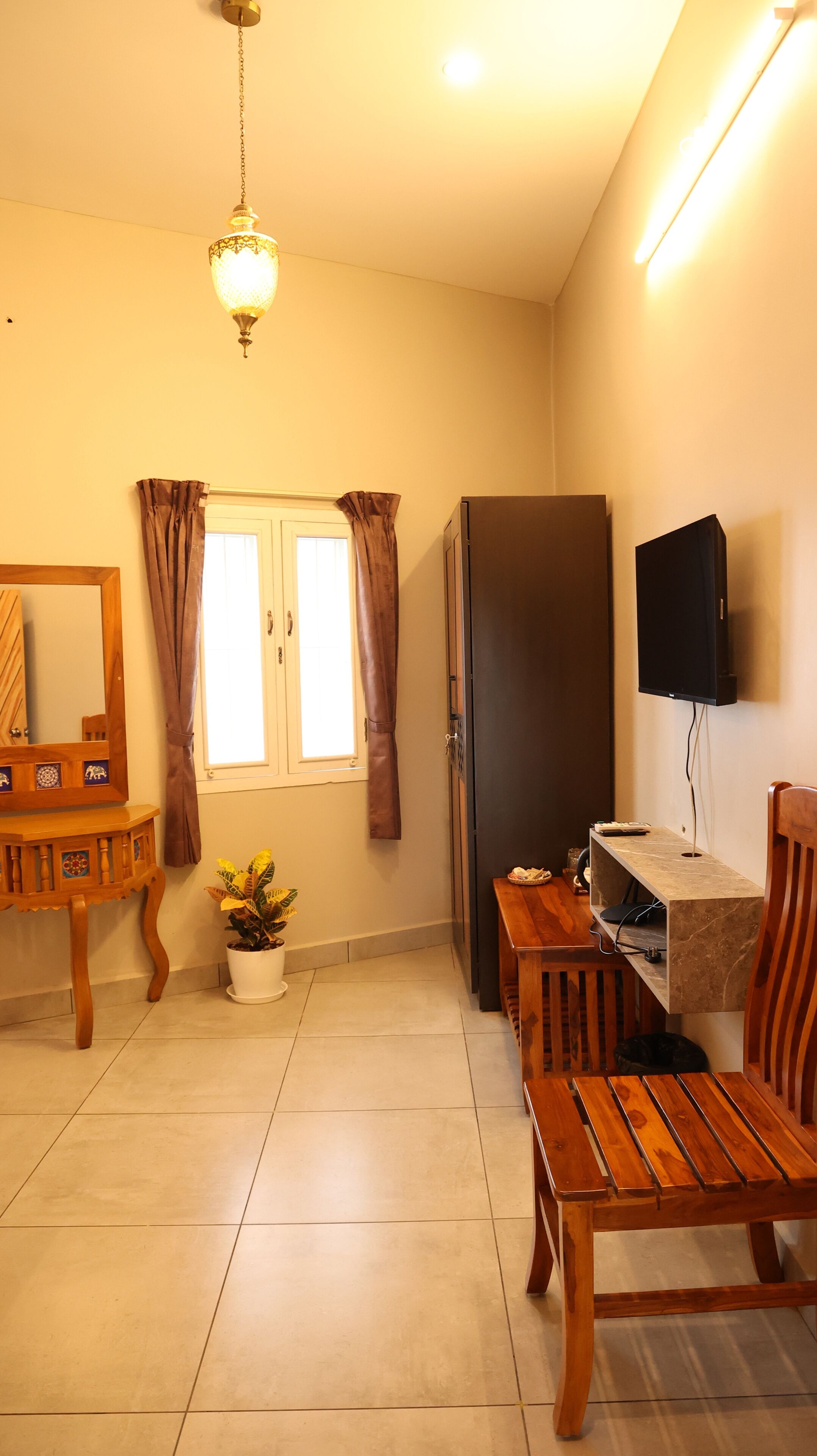 A living room with a wooden coffee table, a TV mounted on the wall, a refrigerator, a potted plant, and a window with curtains.