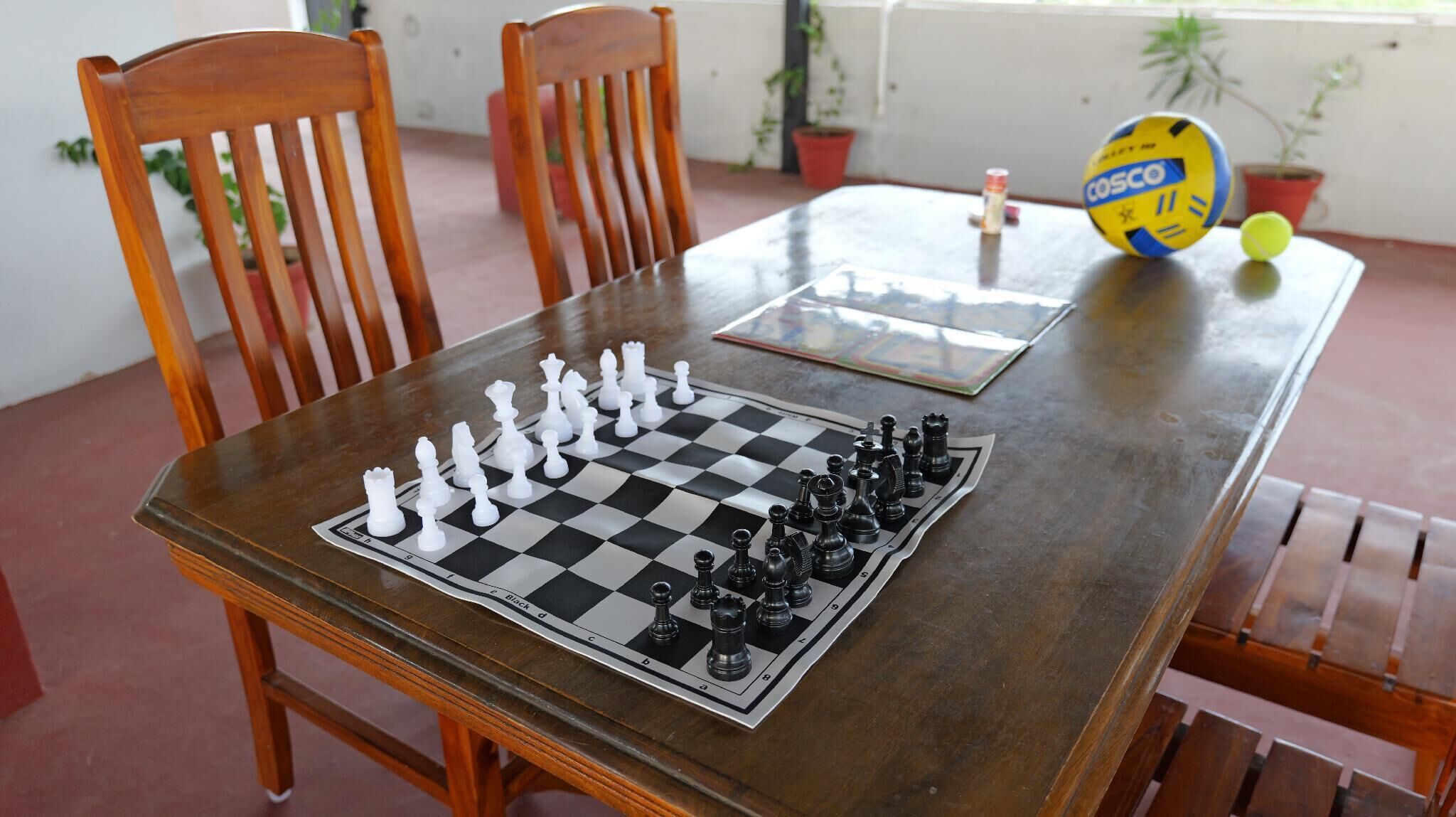A chessboard set on a wooden table with chairs around it.