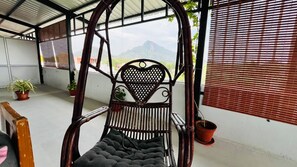A wicker chair with a heart-shaped backrest, a cushioned seat, and a metal frame, positioned on a balcony with potted plants and a view of mountains.