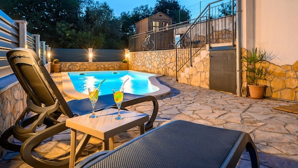 Una piscina exterior con sillas de descanso y una mesa, rodeada de muros de piedra y una escalera.