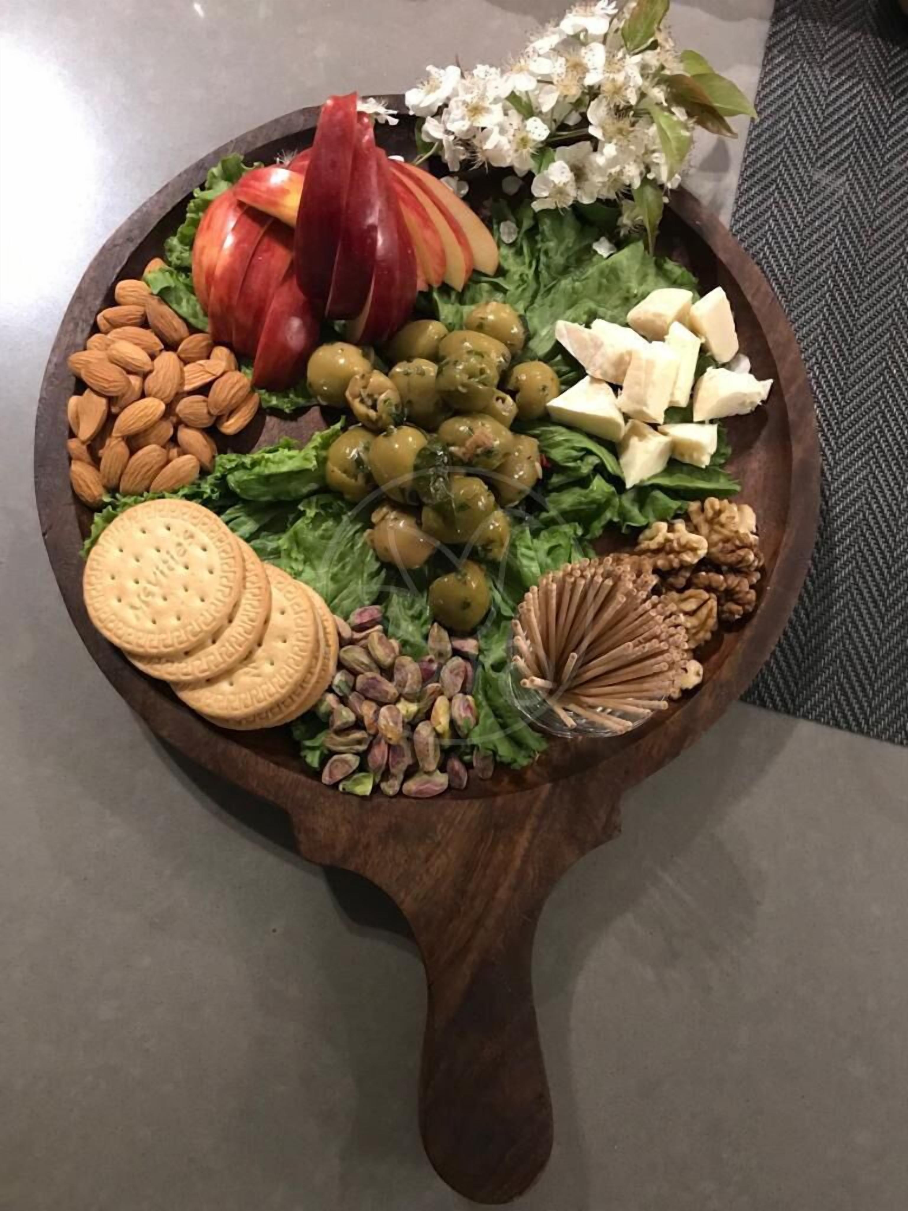 Ein Holzbruch mit verschiedenen Snacks, darunter Mandeln, Oliven, Walnüsse, Crackern sowie einer dekorativen Anordnung aus Blumen und Früchten.