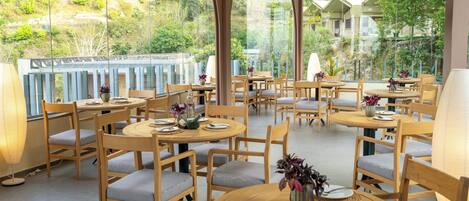 A restaurant with round tables and chairs, set against a backdrop of greenery and buildings.
