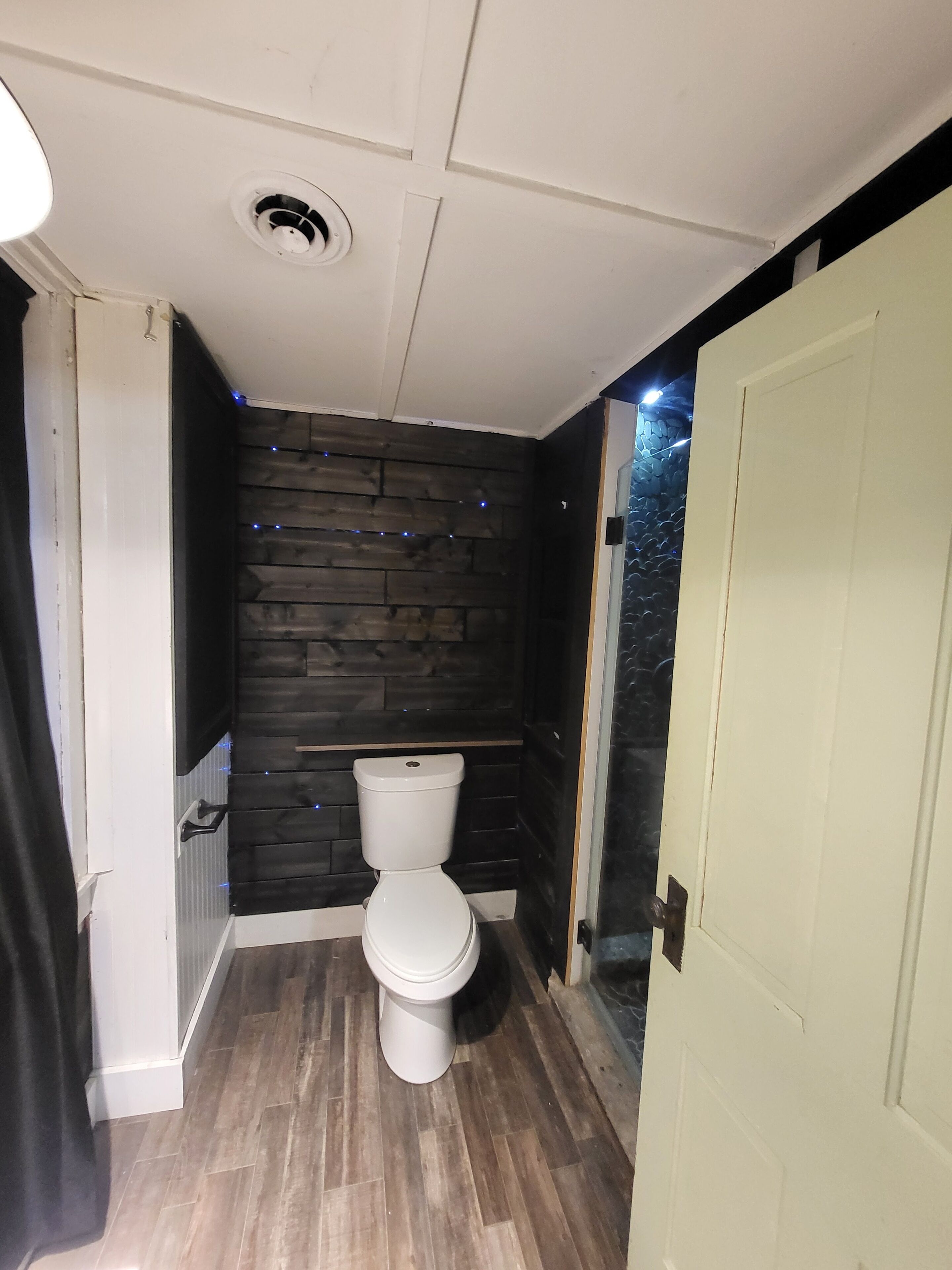 Ein Badezimmer mit Toilette in Weiß, Holzboden, einer dunkel gefärbten Wand mit Struktur und einem Lüftungsgitter an der Decke, sowie einer Tür mit Glasfenster.