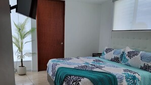 Un dormitorio moderno con una cama, televisor montado en la pared, una planta en maceta y una ventana con persianas.
