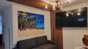 Una sala moderna con un sofá gris, un televisor de pantalla plana grande montado en la pared y un cuadro con una playa tropical.